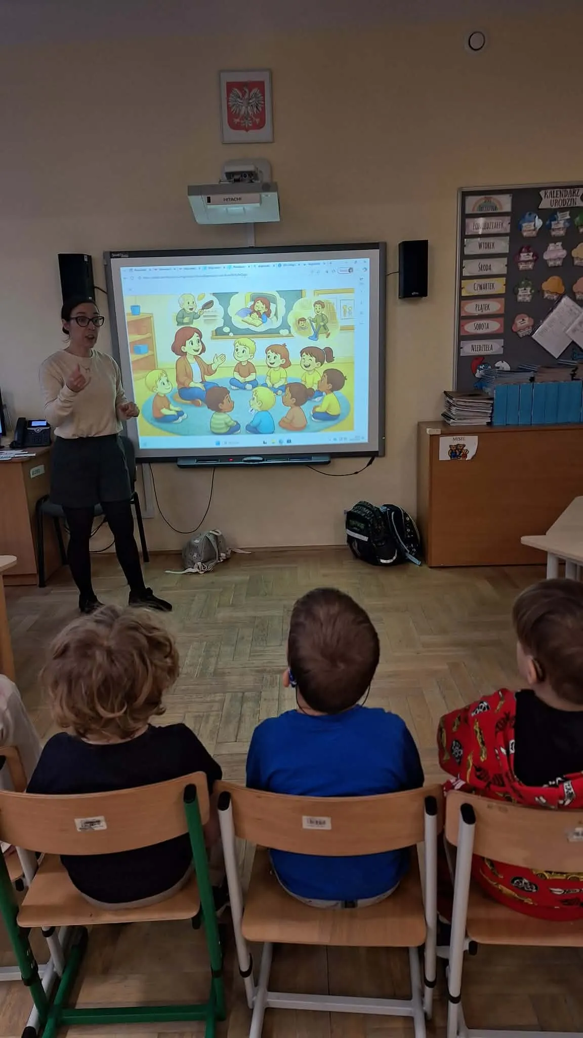 Nauczycielka stoi przy tablicy interaktywnej i prowadzi zajęcia dla siedzących dzieci.
