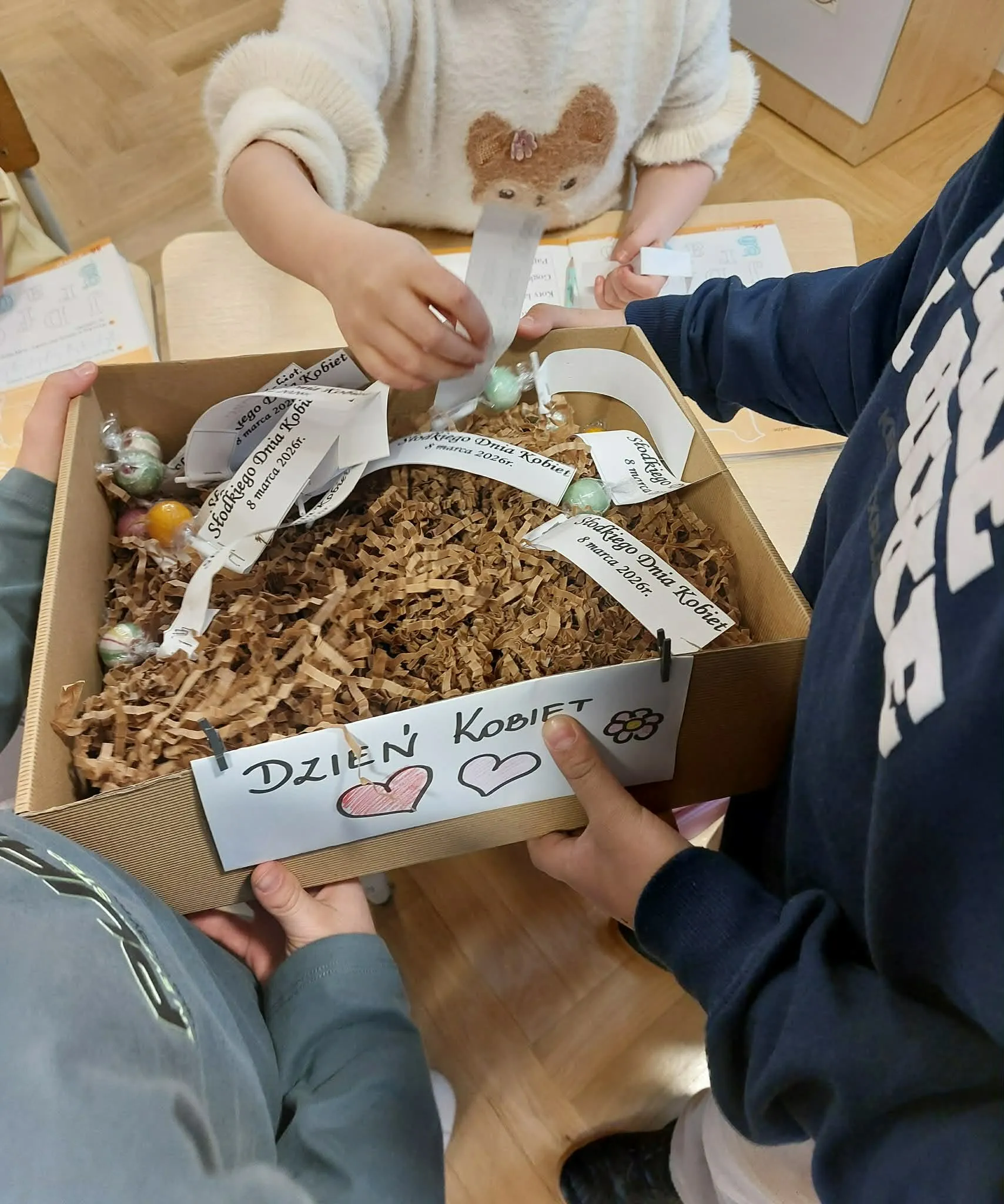 Dzieci losują z pudełka karteczki z życzeniami i słodkie upominki z okazji Dnia Kobiet.