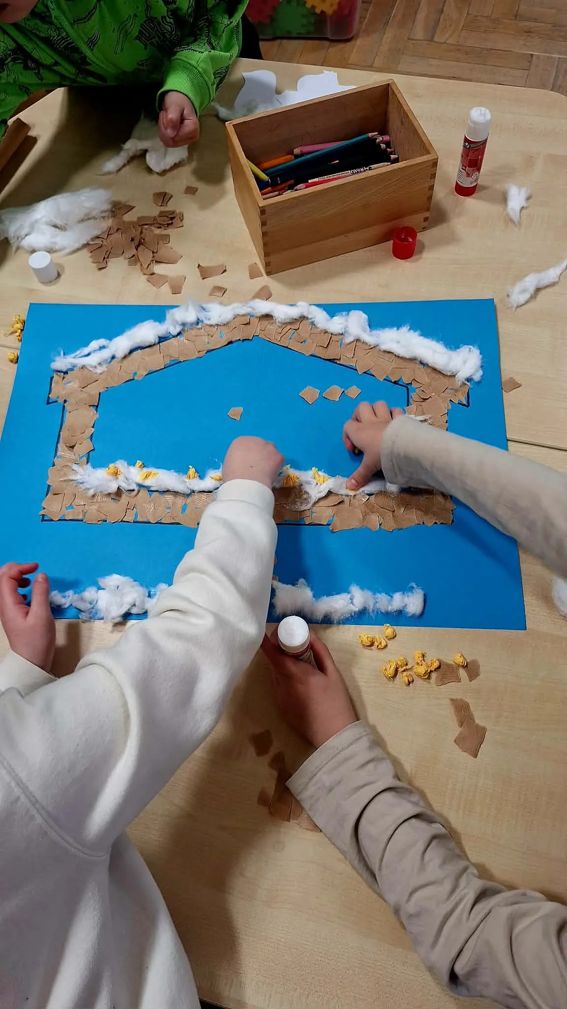 Dzieci przyklejają watę i kawałki papieru, tworząc pracę plastyczną z ptakami na niebieskim tle.