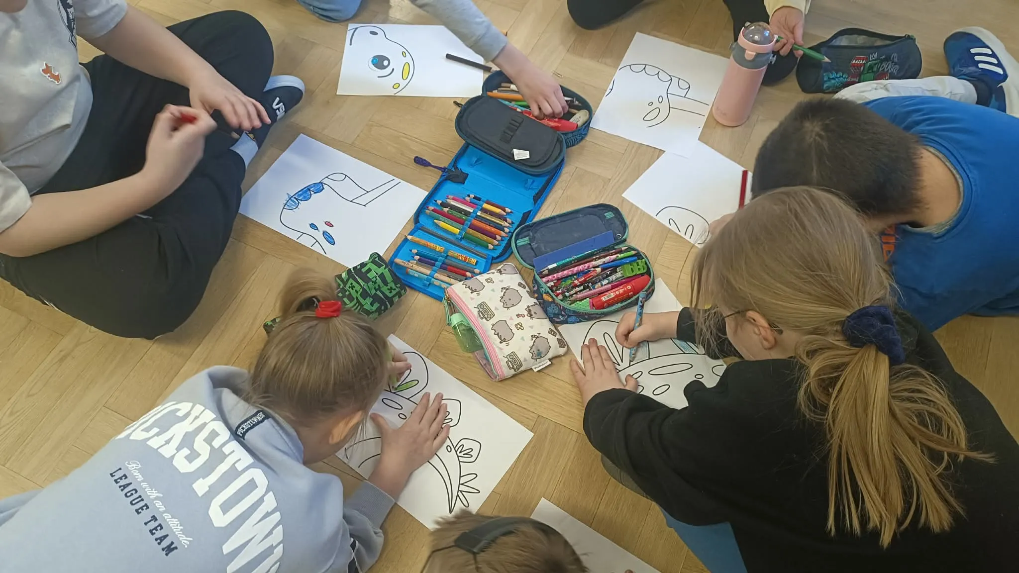 Uczniowie siedzą na podłodze i kolorują rysunki dinozaurów, korzystając z kredek i piórników.