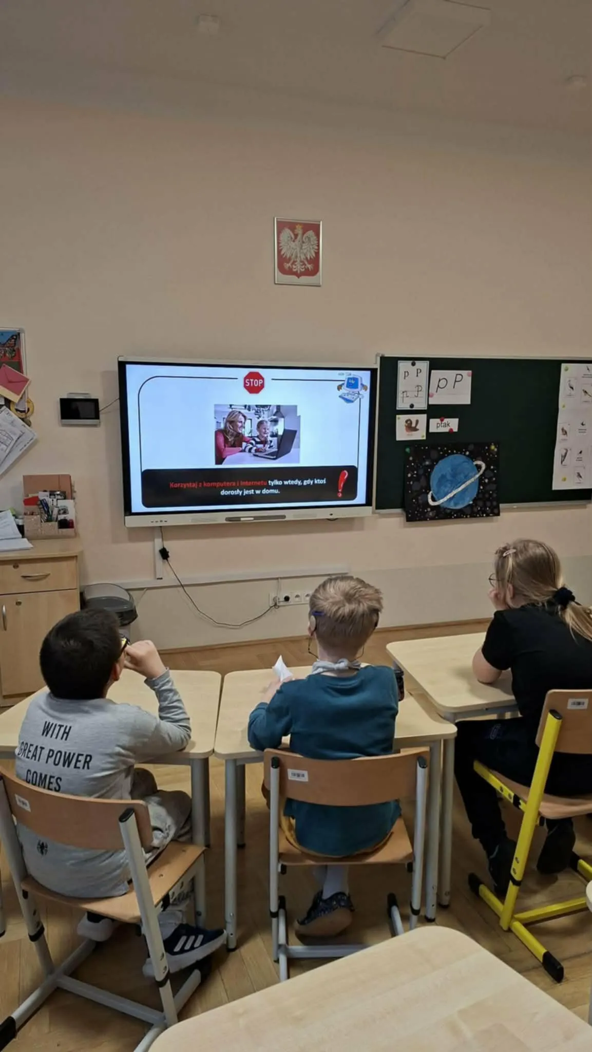 Troje uczniów siedzi w ławkach i ogląda na ekranie prezentację dotyczącą bezpiecznego korzystania z internetu.