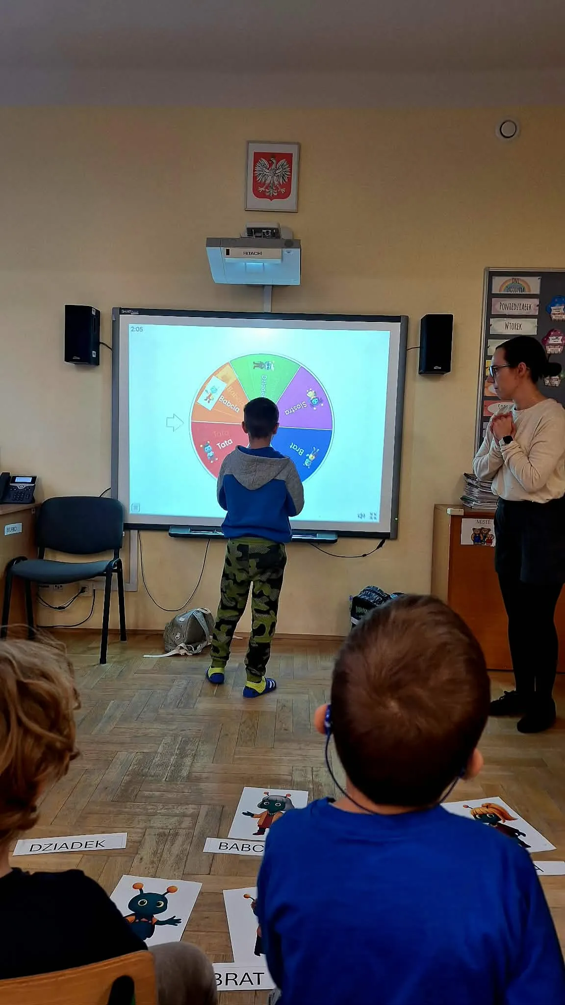 Chłopiec stoi przy tablicy interaktywnej z kołem wyboru, a nauczycielka obserwuje.
