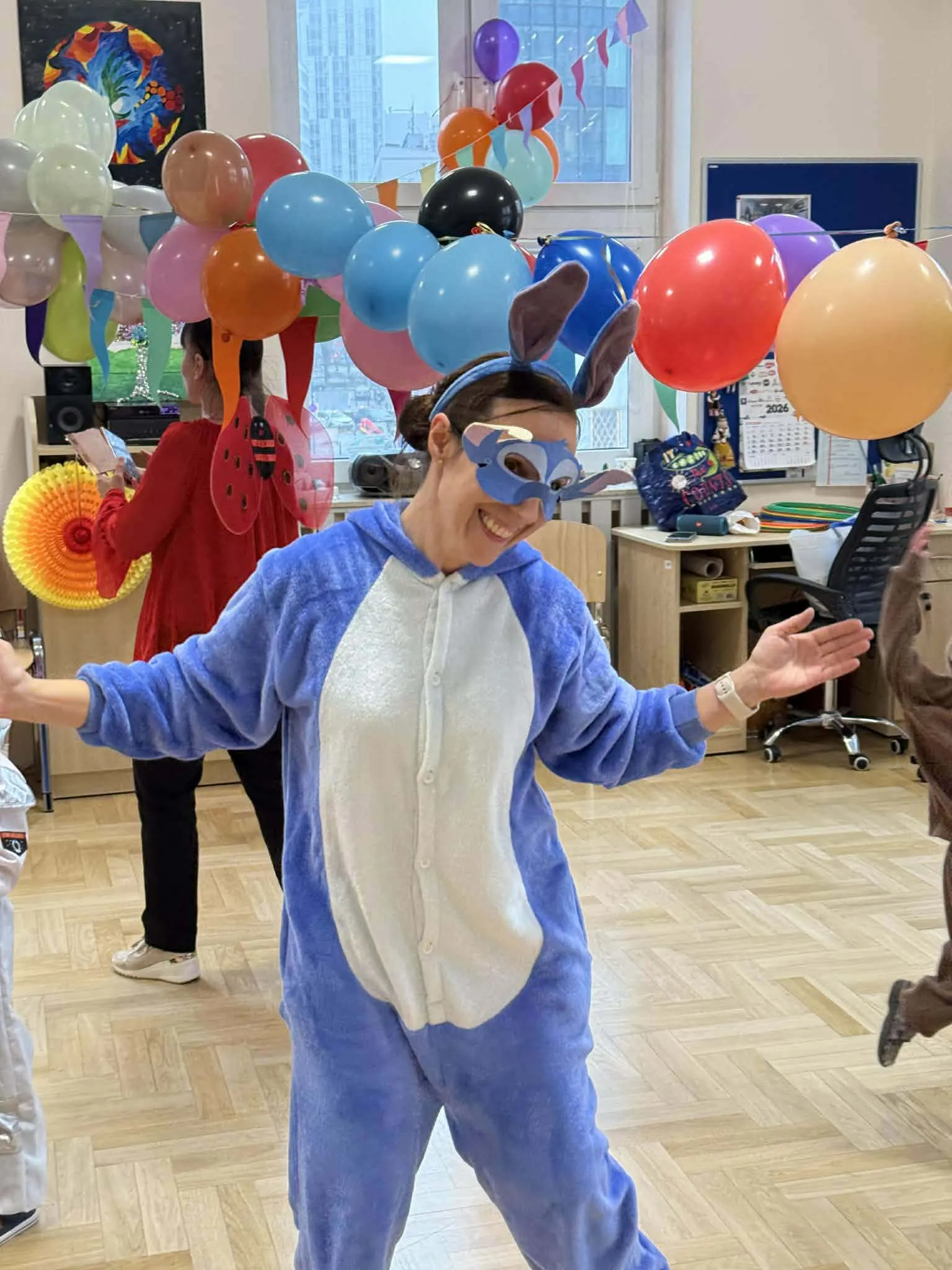 Uśmiechnięty animator w niebieskim kostiumie tańczy wśród balonów podczas szkolnej zabawy.