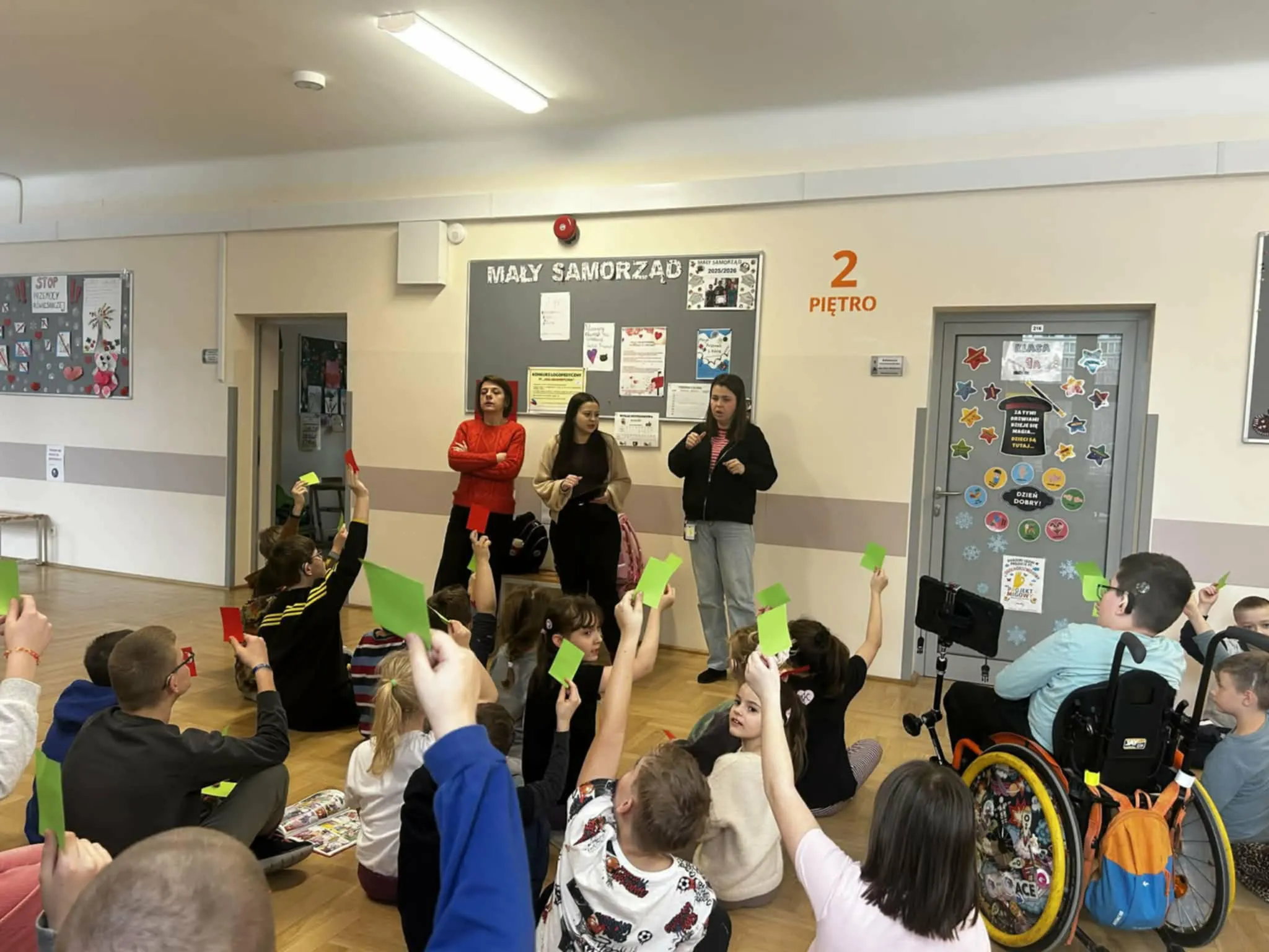 Uczniowie siedzą na podłodze i podnoszą zielone oraz czerwone kartki podczas zajęć prowadzonych przez nauczycielki.