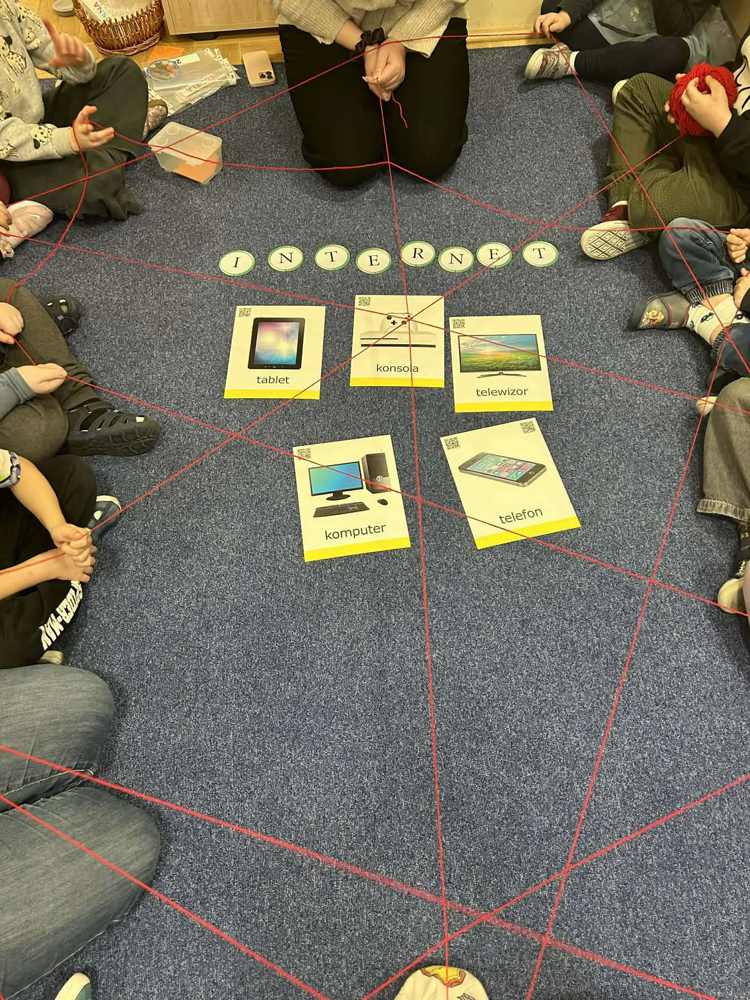 Dzieci siedzą w kręgu na dywanie i tworzą sieć z czerwonej włóczki wokół kart z urządzeniami podłączonymi do internetu.