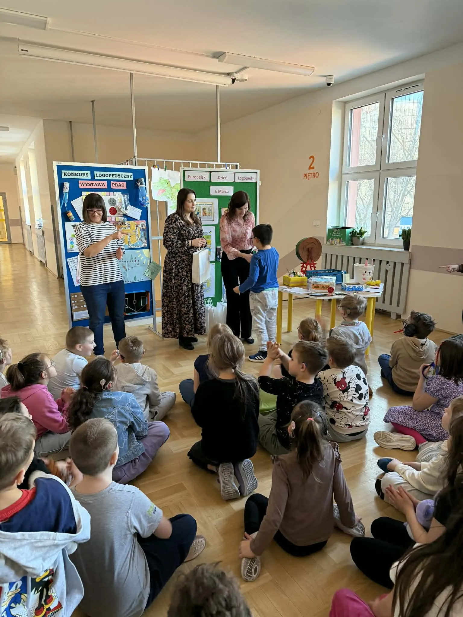Nauczycielki wręczają nagrodę chłopcu, a dzieci siedzą na podłodze i obserwują wydarzenie