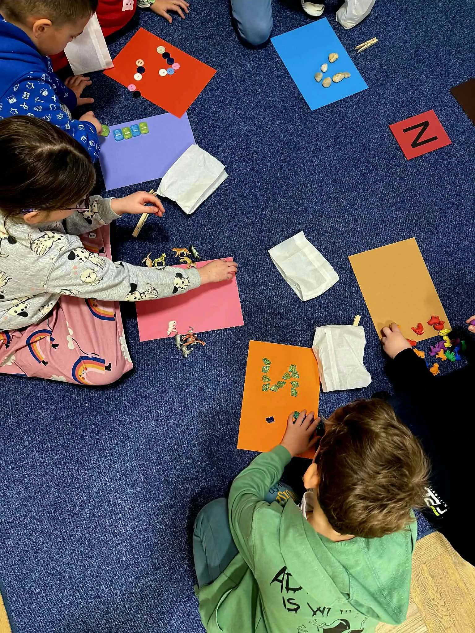 Dzieci siedzą na dywanie i układają litery oraz elementy na kolorowych kartkach podczas zajęć edukacyjnych.