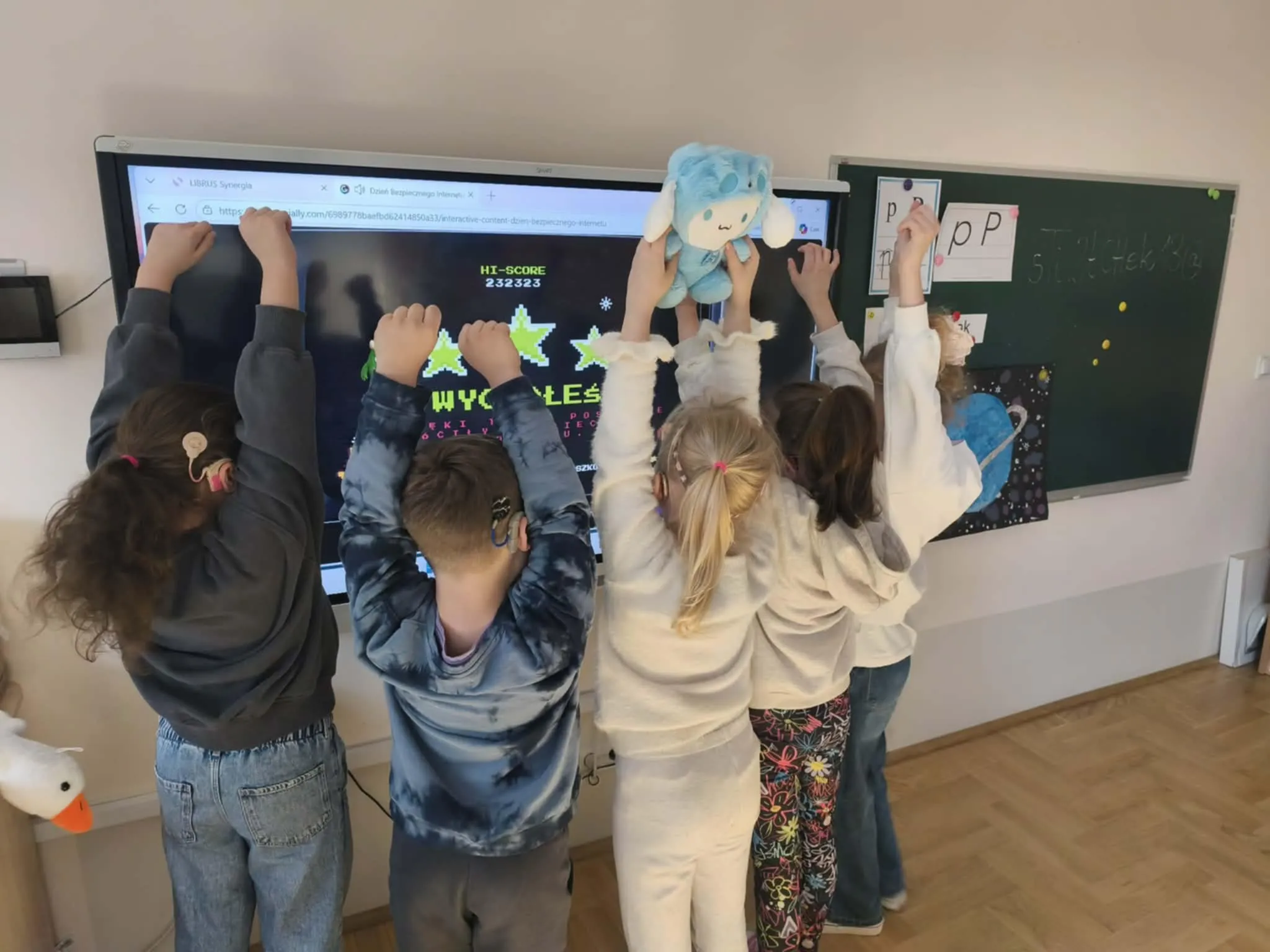 Uczniowie stoją przed tablicą interaktywną i unoszą ręce podczas wspólnej aktywności w klasie.