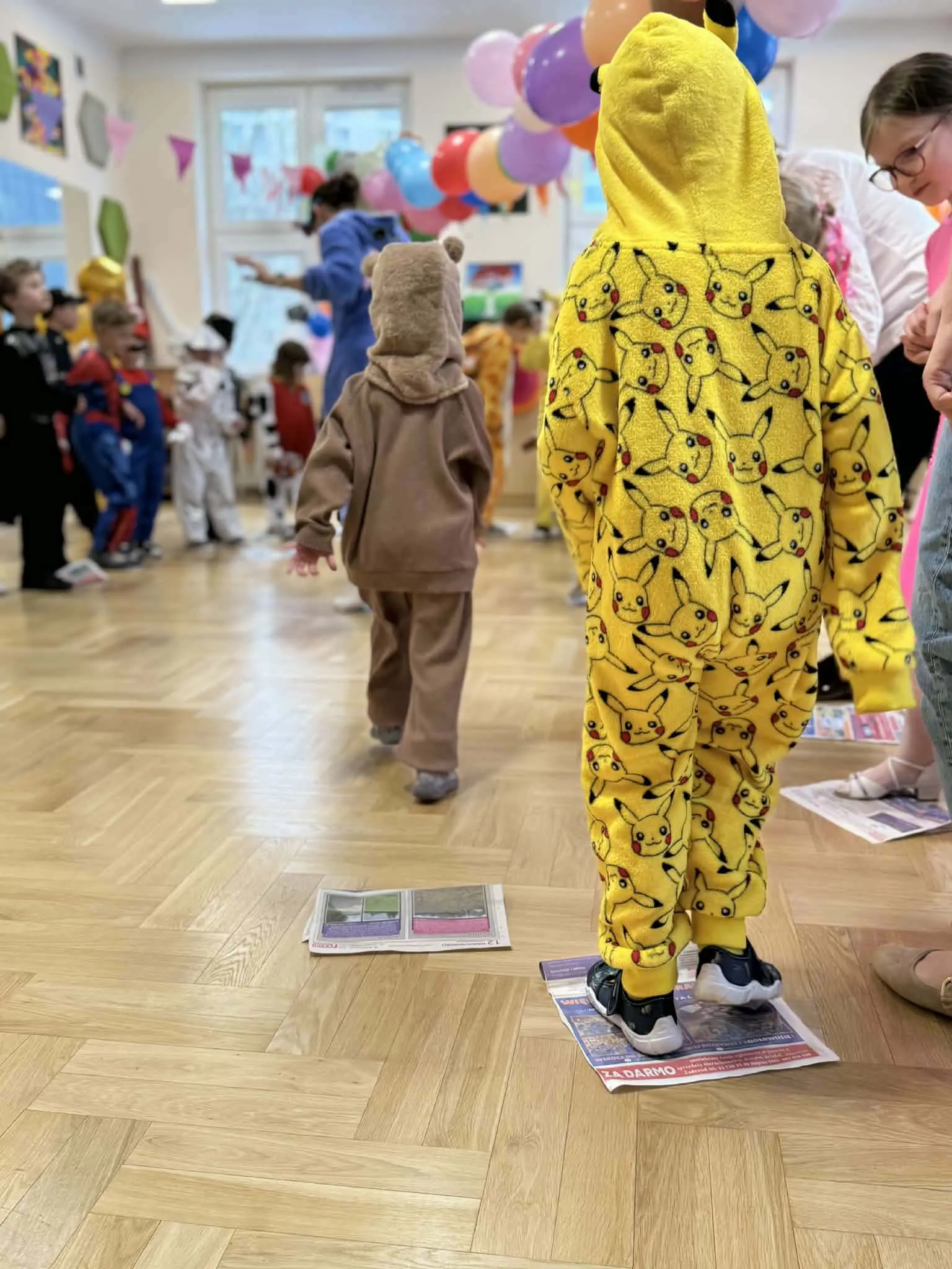 Dzieci w kolorowych kostiumach biorą udział w zabawie karnawałowej w sali udekorowanej balonami.