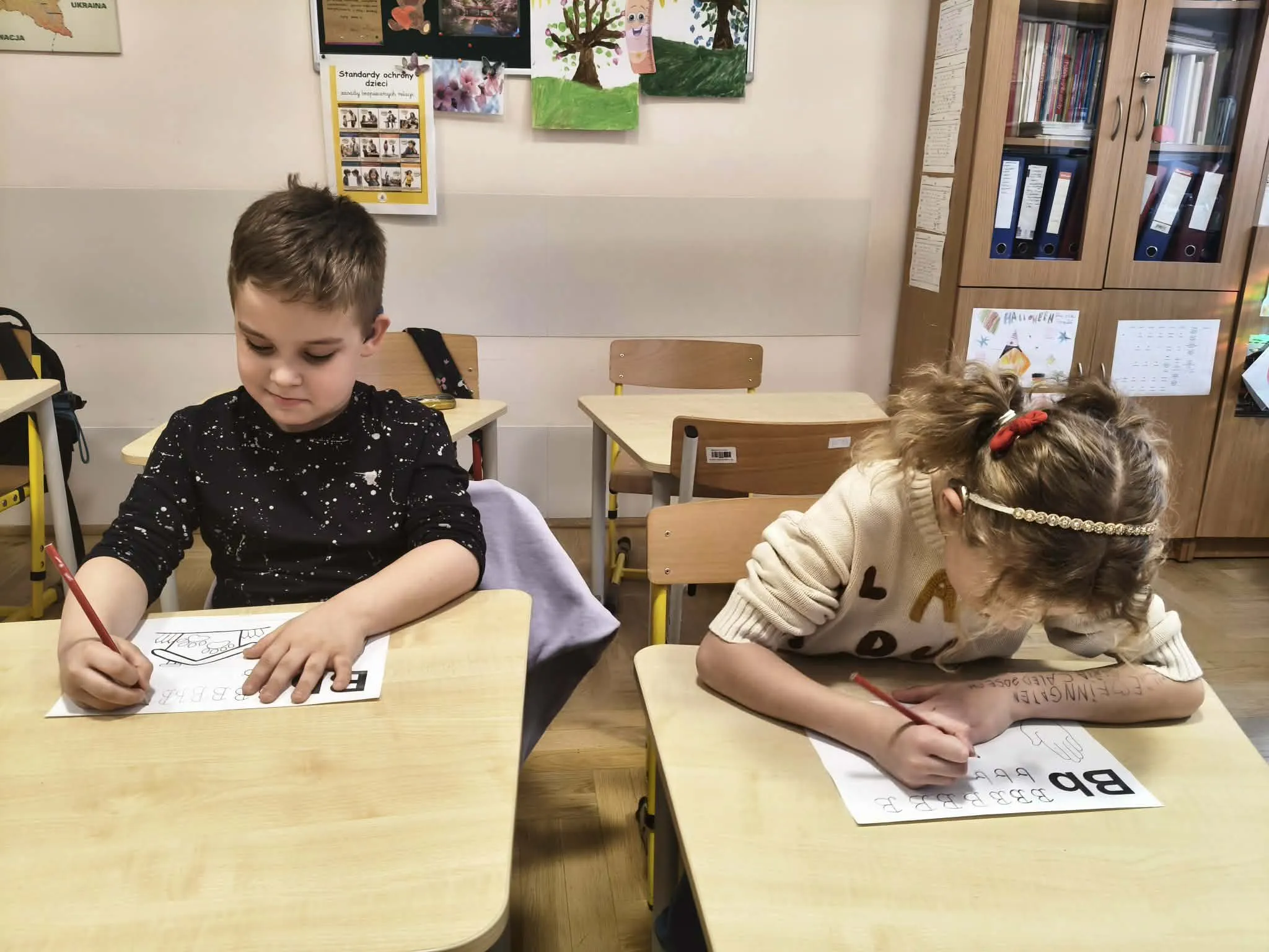 Dwoje dzieci ćwiczy pisanie liter przy szkolnych ławkach