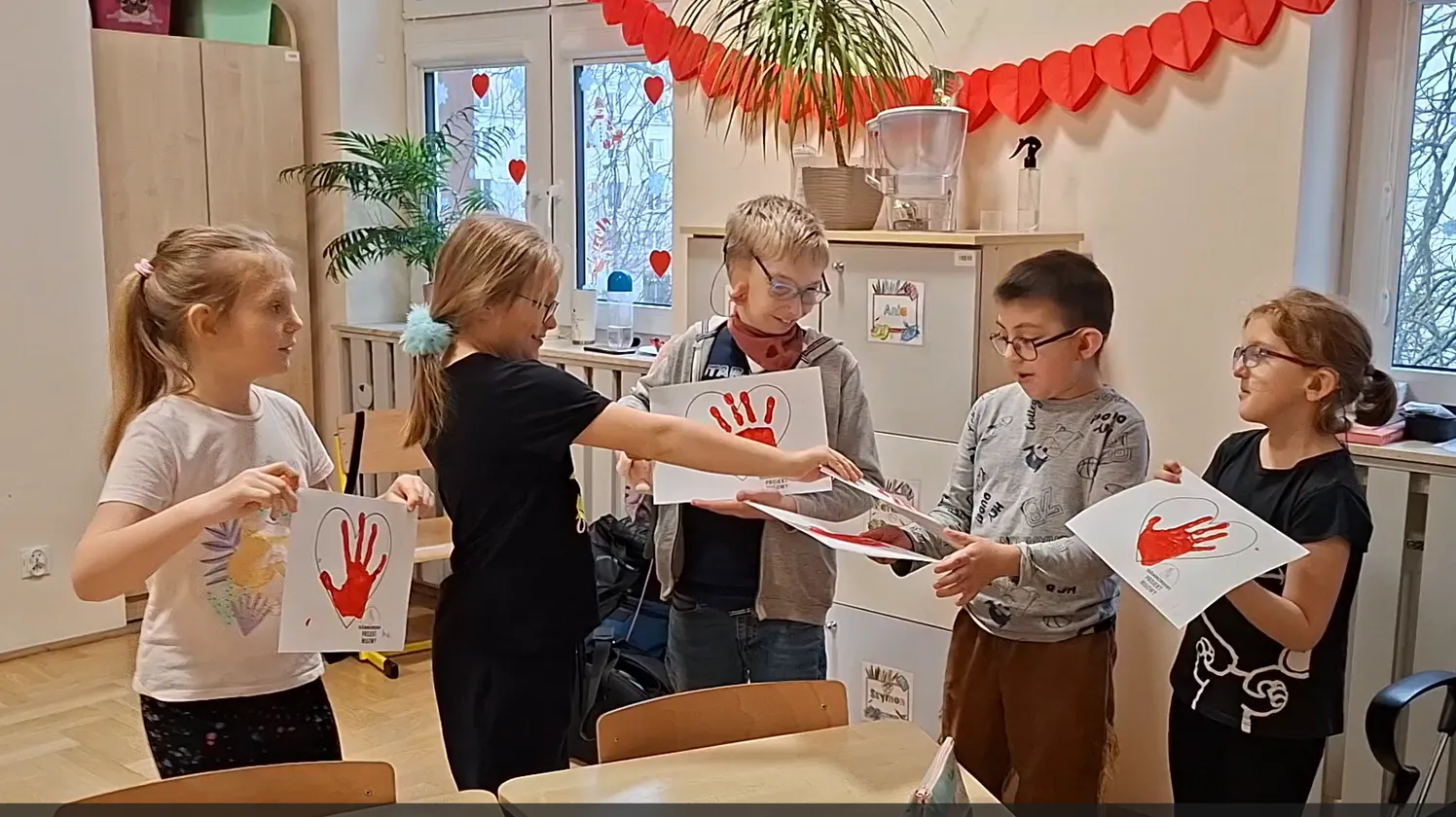 Dzieci w klasie pokazują prace plastyczne z czerwonymi odciskami dłoni