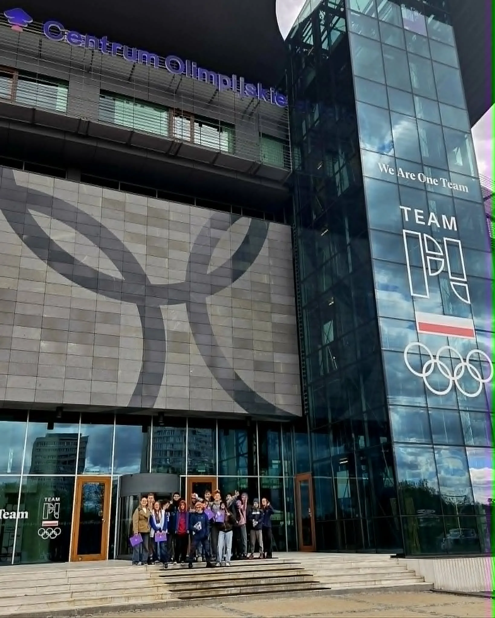 Grupa uczniów stoi przed wejściem do Centrum Olimpijskiego w Warszawie podczas wycieczki szkolnej.