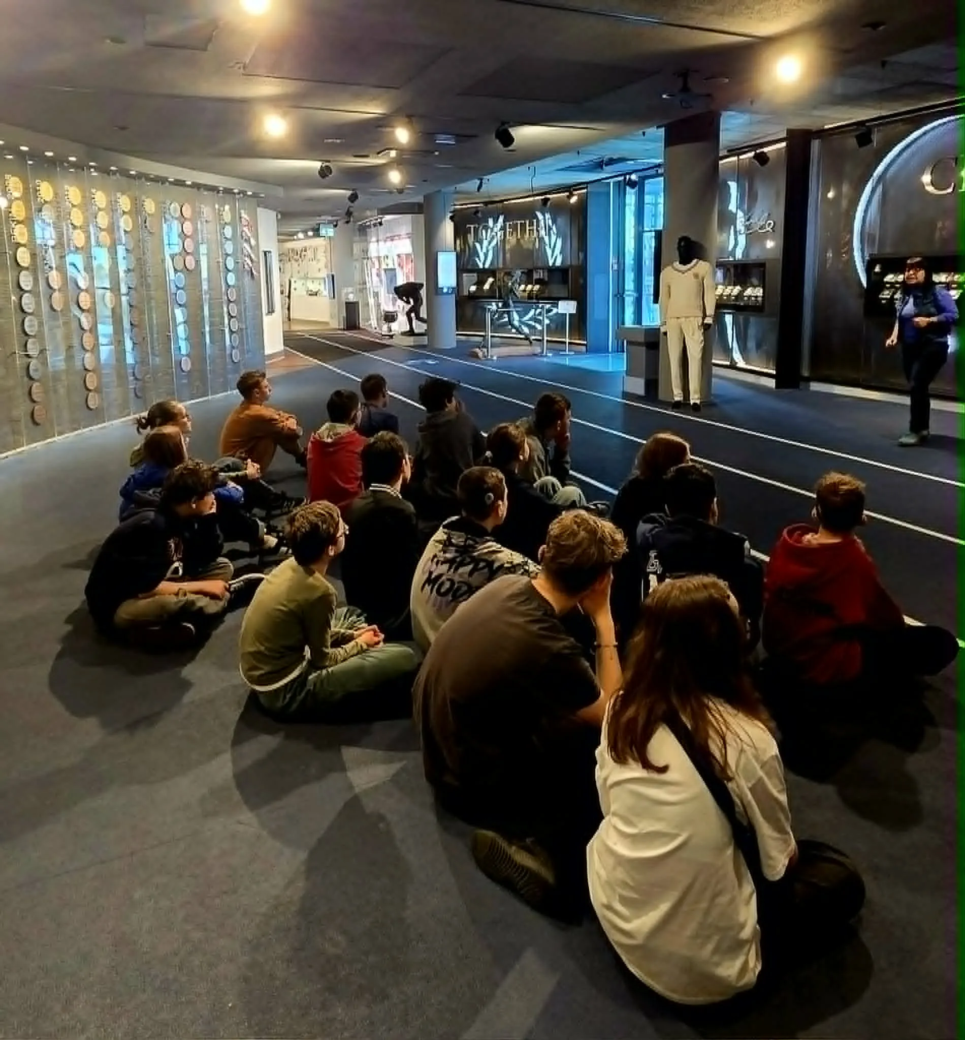 Uczniowie siedzą na podłodze i słuchają przewodnika oprowadzającego po muzeum sportu.