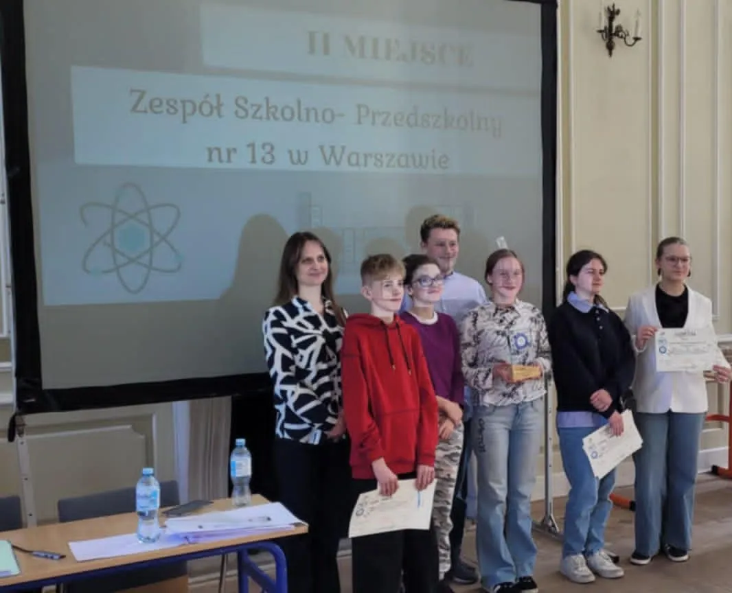 Uczniowie Zespołu Szkolno-Przedszkolnego nr 13 w Warszawie odbierają nagrodę za zajęcie II miejsca podczas konkursu. Grupa stoi na scenie przed ekranem z nazwą szkoły, trzymając dyplomy.