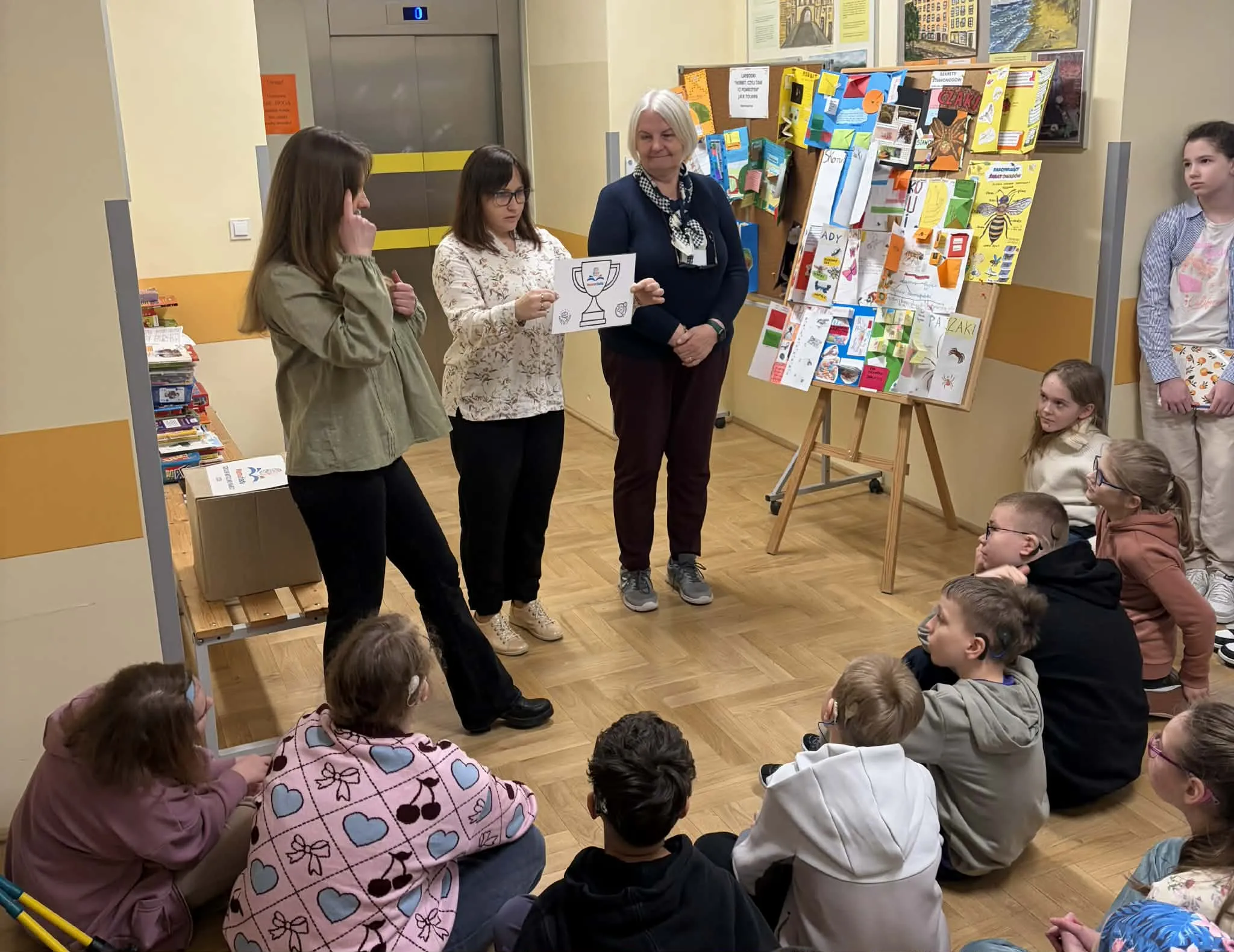 Nauczycielka pokazuje rysunek pucharu na kartce uczniom siedzącym na podłodze