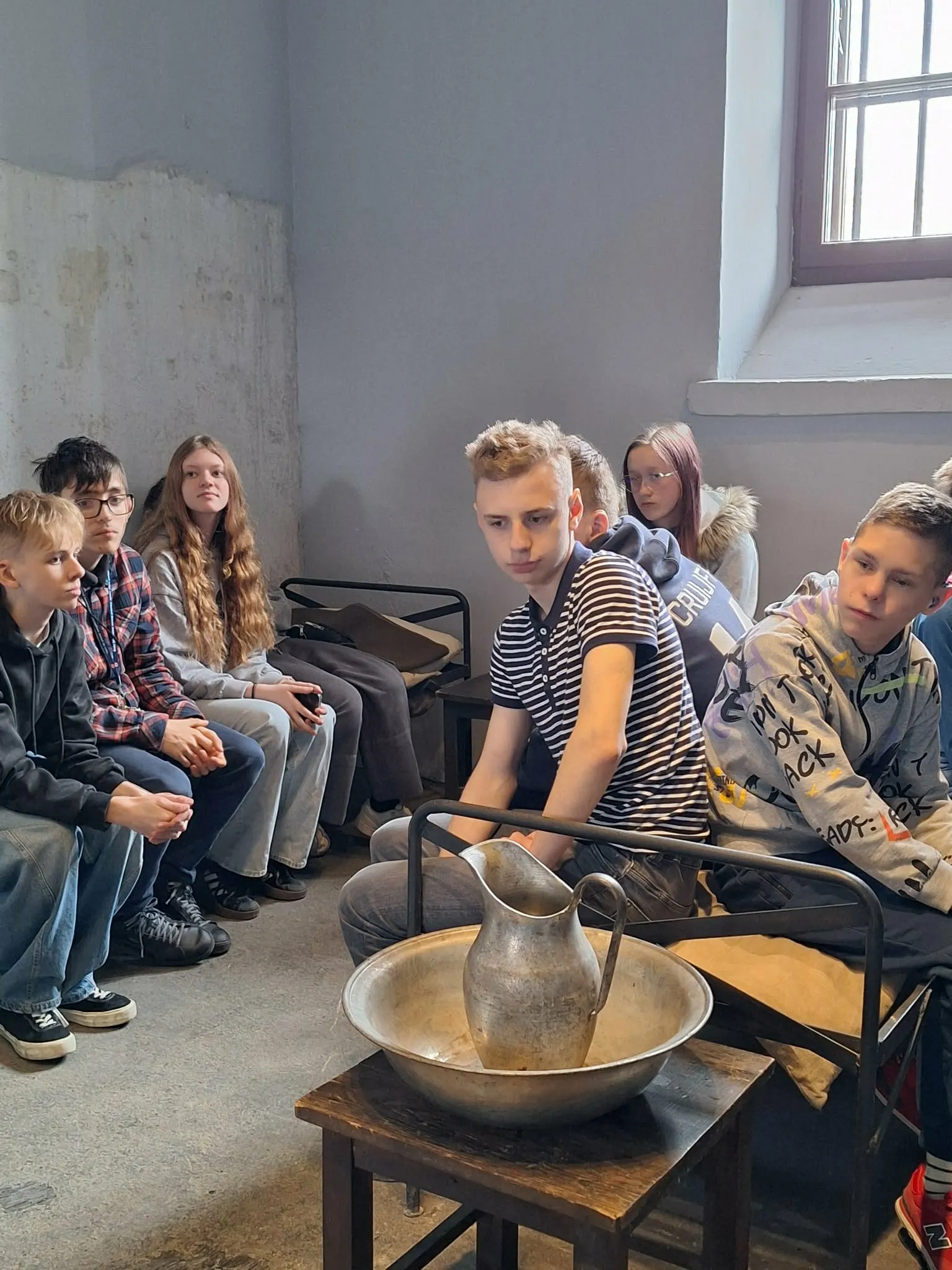 Grupa uczniów siedzi w historycznej celi więziennej podczas zwiedzania muzeum