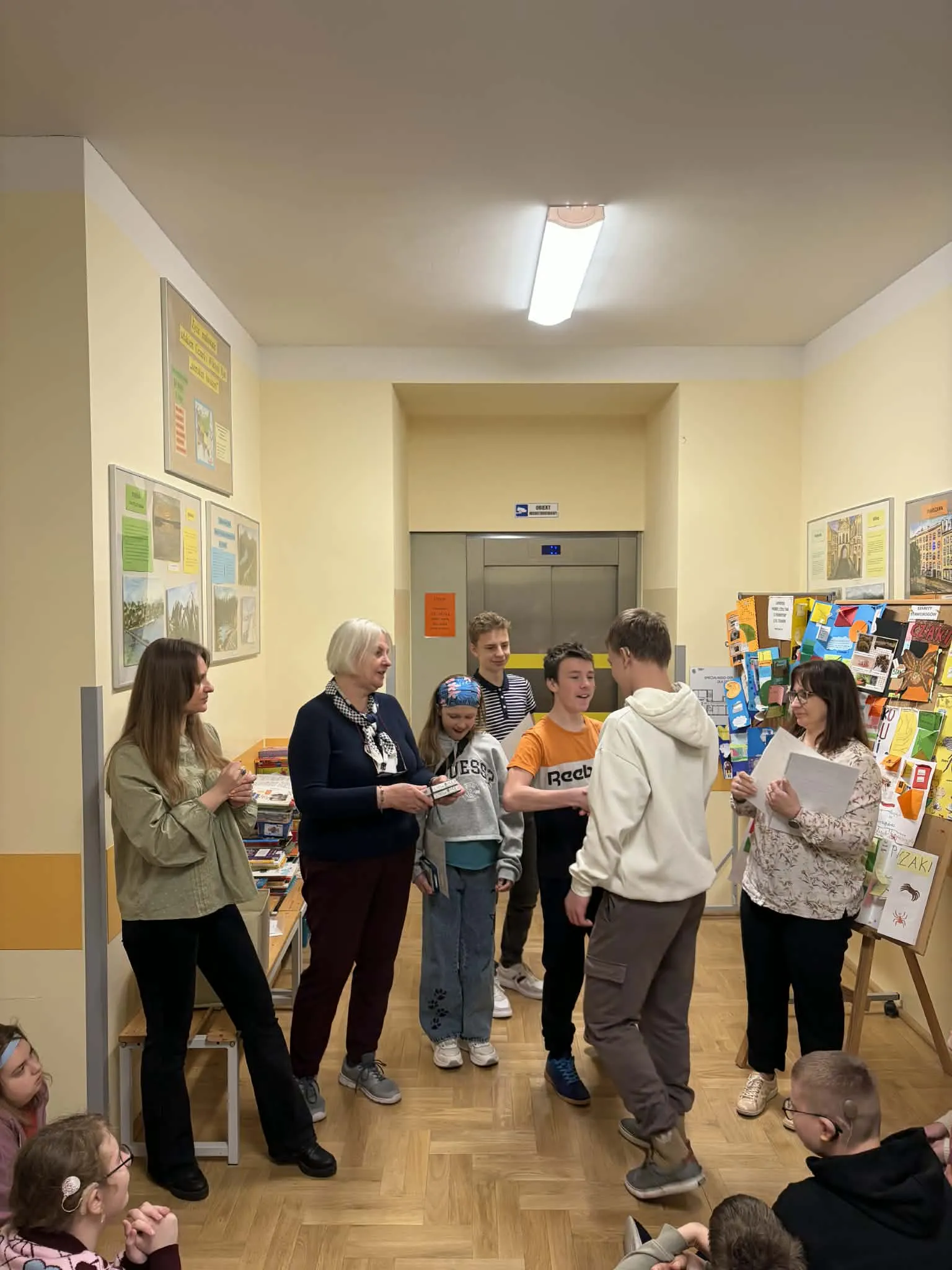 Uczniowie i nauczyciele stoją razem podczas wręczania nagród na szkolnym korytarzu