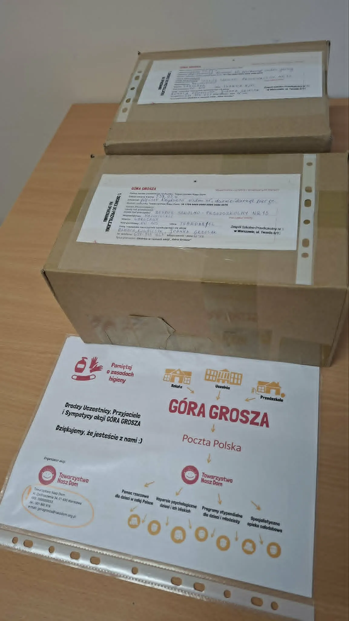 Dwa kartonowe pudełka z zapakowanymi monetami oraz plakat akcji charytatywnej Góra Grosza ustawione na szkolnym stoliku.