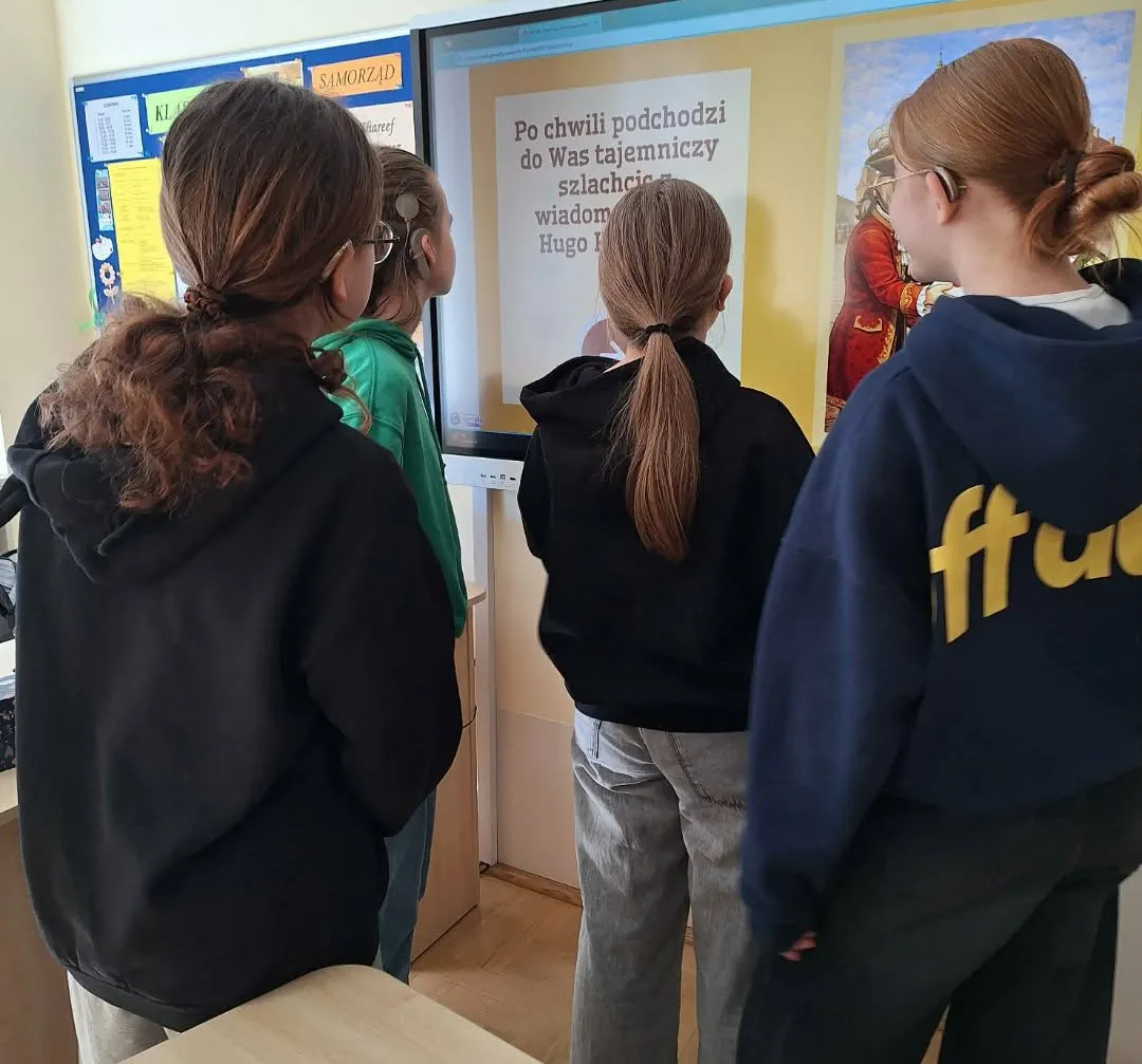 Grupa uczennic stoi przed tablicą multimedialną i uczestniczy w zadaniu dotyczącym historii Polski.