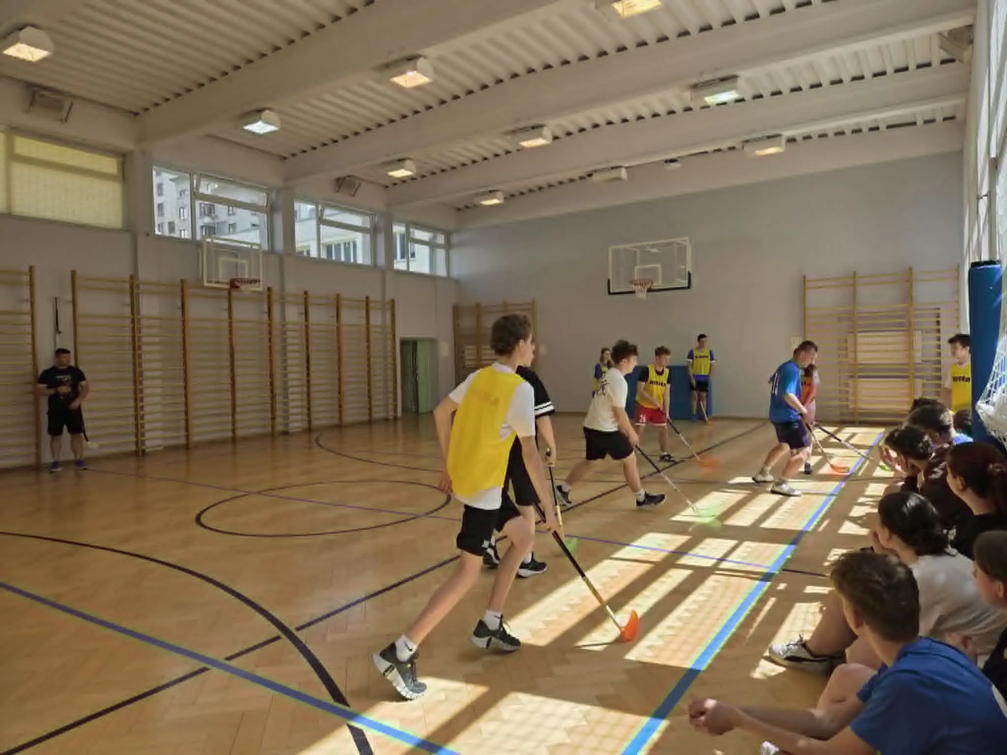 Zawodnicy walczą o piłeczkę podczas meczu unihokeja rozgrywanego w szkolnej sali sportowej.