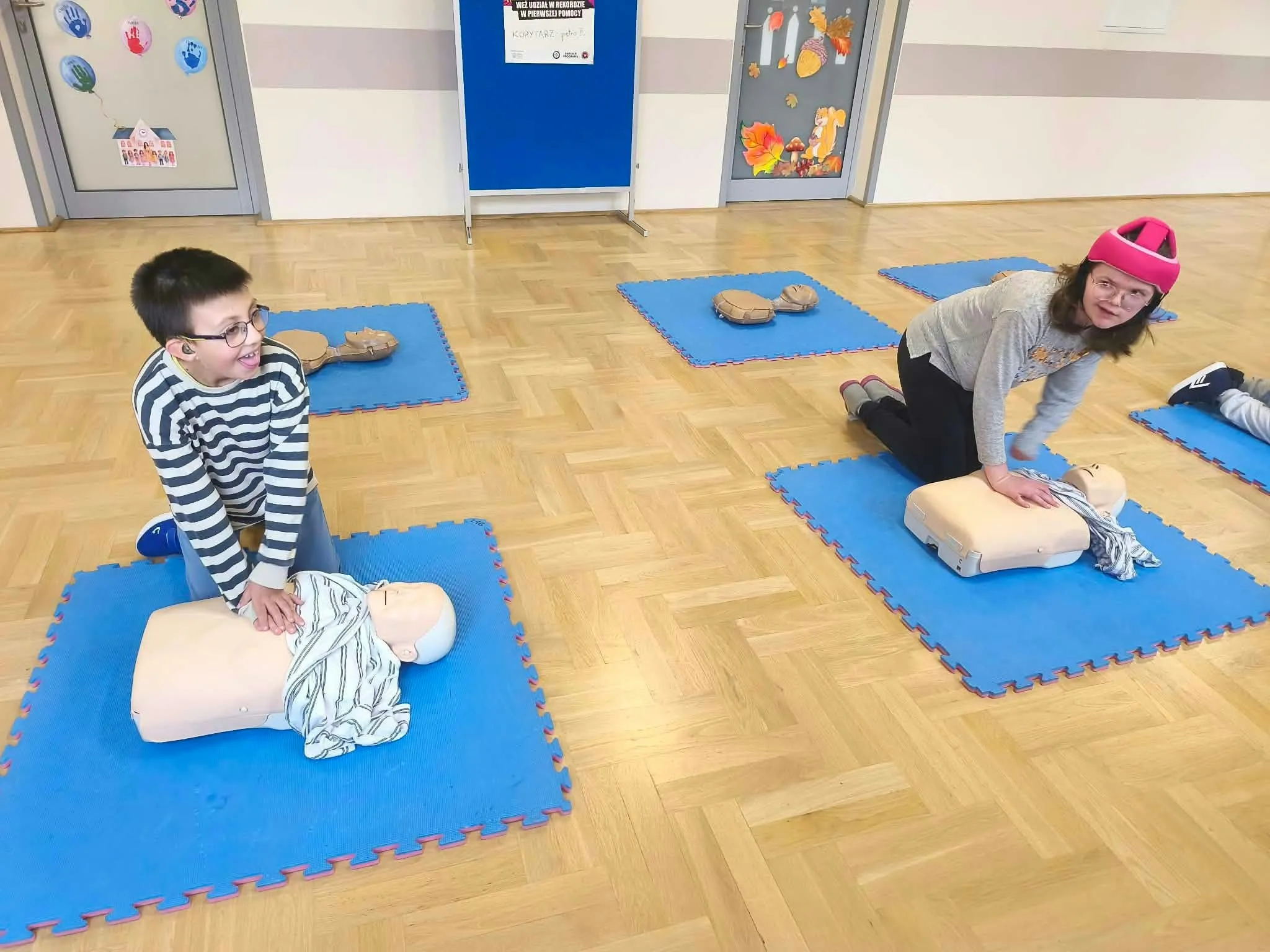 Dwoje uczniów ćwiczy uciskanie klatki piersiowej na fantomach do resuscytacji, korzystając z niebieskich mat rozłożonych na podłodze.