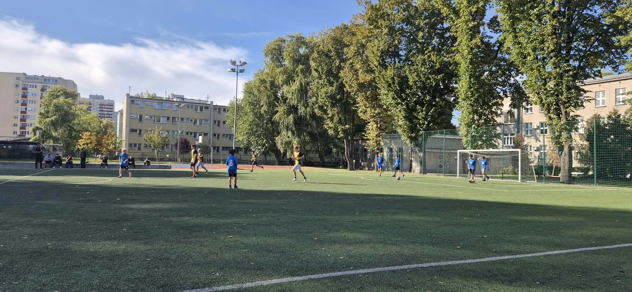 Uczniowie podczas meczu piłki nożnej na szkolnym boisku. Zawodnicy dwóch drużyn rywalizują ze sobą podczas rozgrywki.