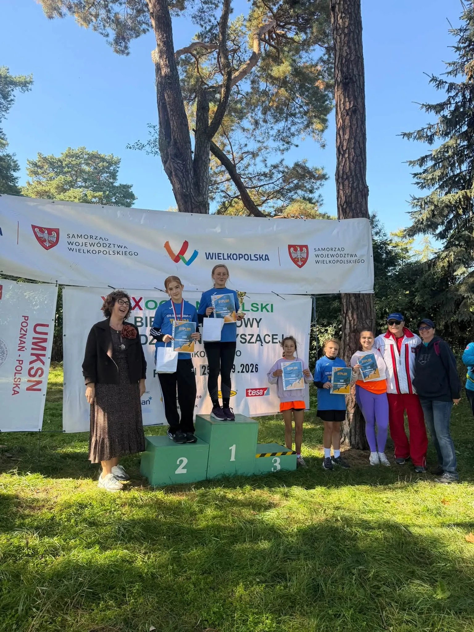 Zawodniczki podczas ceremonii dekoracji na podium XXIX Ogólnopolskich Biegów Przełajowych Młodzieży Niesłyszącej w Poznaniu. Uczestniczki prezentują medale, puchar i dyplomy.