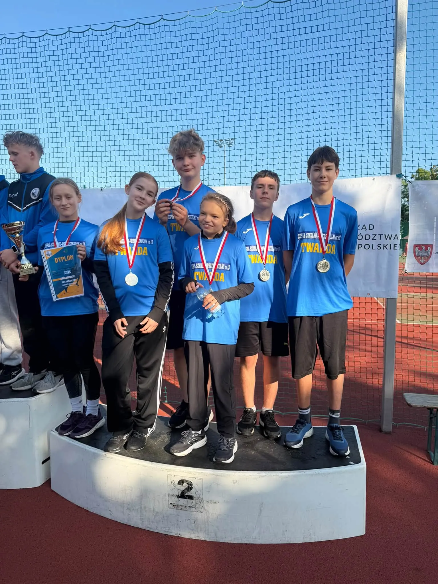 Reprezentacja ZSP nr 13 na podium po zdobyciu drugiego miejsca podczas XXIX Ogólnopolskich Biegów Przełajowych Młodzieży Niesłyszącej w Poznaniu. Uczniowie prezentują zdobyte medale i dyplom.