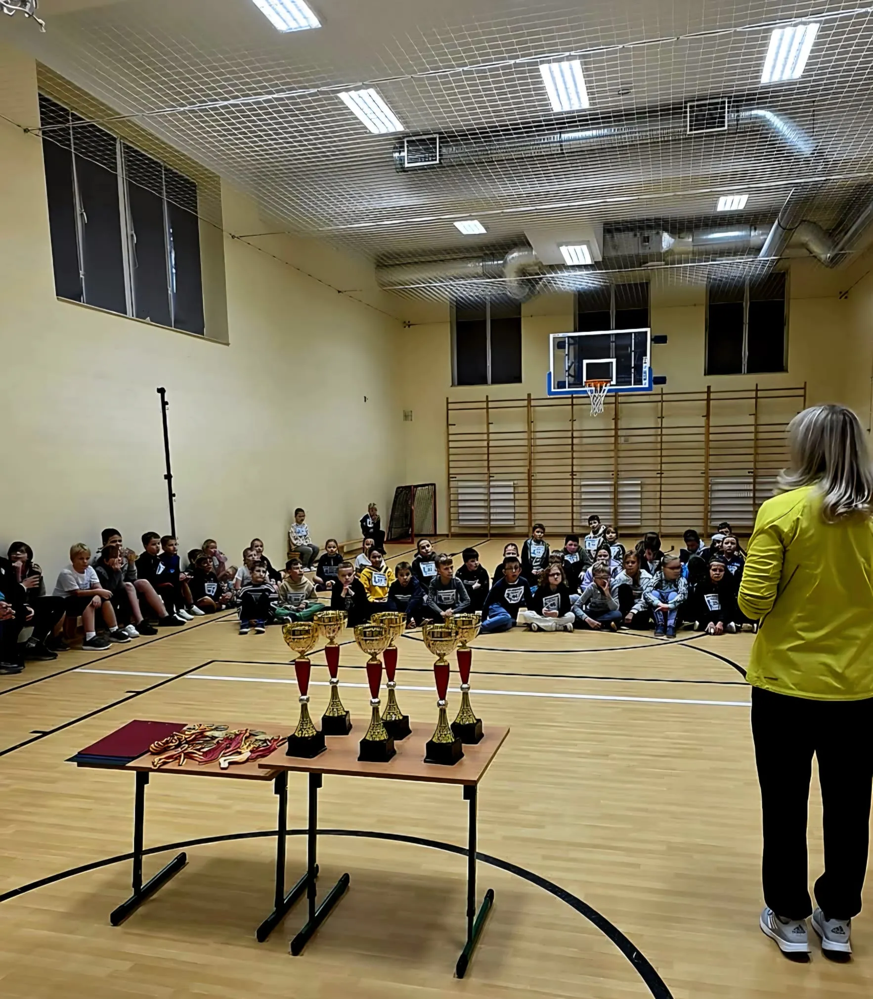 Uczniowie zgromadzeni w sali gimnastycznej podczas podsumowania zawodów lekkoatletycznych. Na stole przygotowano puchary i medale dla uczestników.