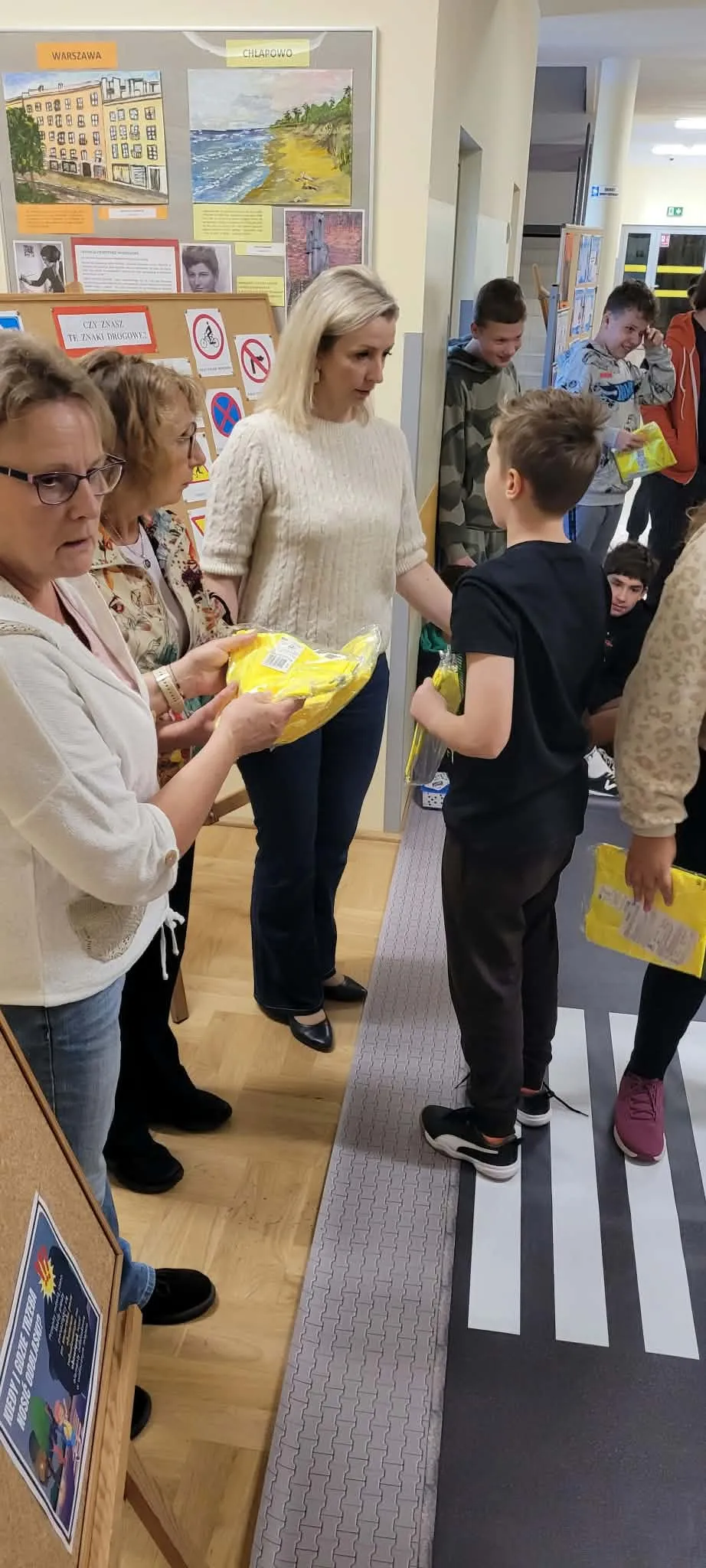 Uczniowie wraz z nauczycielami uczestniczą w zajęciach dotyczących bezpieczeństwa na drodze, prowadzonych przy przygotowanym przejściu dla pieszych i tablicy ze znakami drogowymi.